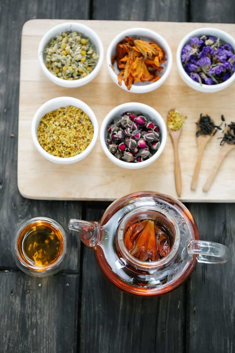 A variety of dried flowers and herbal tea in bowls on a wooden surface, captured in a flat lay composition.