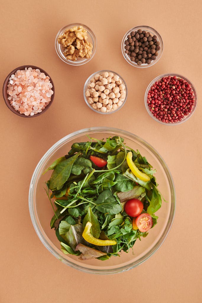 A vibrant salad with fresh greens, peppers, and tomatoes alongside bowls of spices and nuts, captured from above.