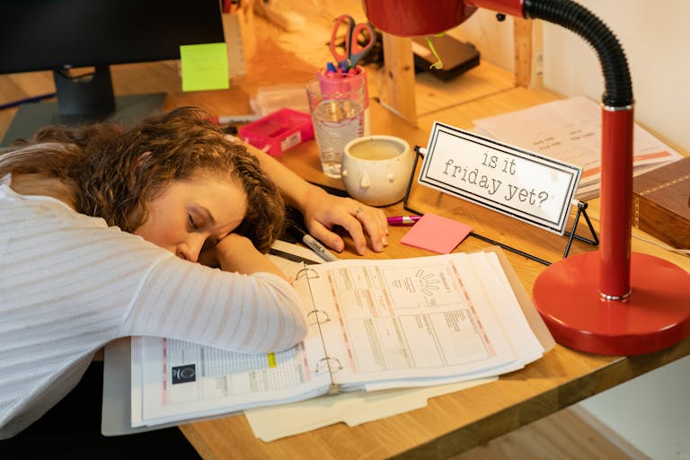 A woman sleeping at her desk with office documents, a coffee cup, and a red desk lamp.